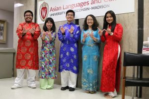 Five people wearing colorful traditional Vietnamese attire smile and form a W with their fingers in front of a University of Wisconsin-Madison sign. 
