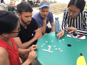 Four people sit around a green table outdoors, focused on a word game with letter tiles. 