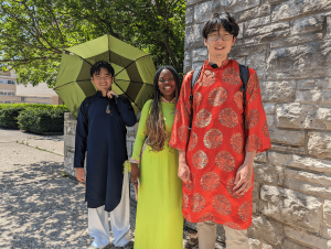 Three people in vibrant clothing stand outdoors; one holds a green umbrella.