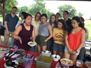 A group stands in front of a table of food. One person is pointing and talking about the food. 