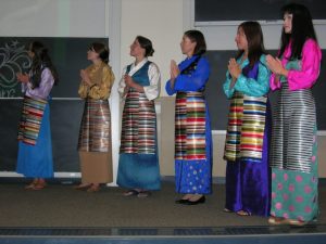 Six women stand in a line wearing colorful traditional outfits and aprons with striped patterns. They are clasping their hands in front of a classroom setting.