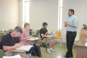 A teacher stands at the front of a classroom speaking to three seated students.