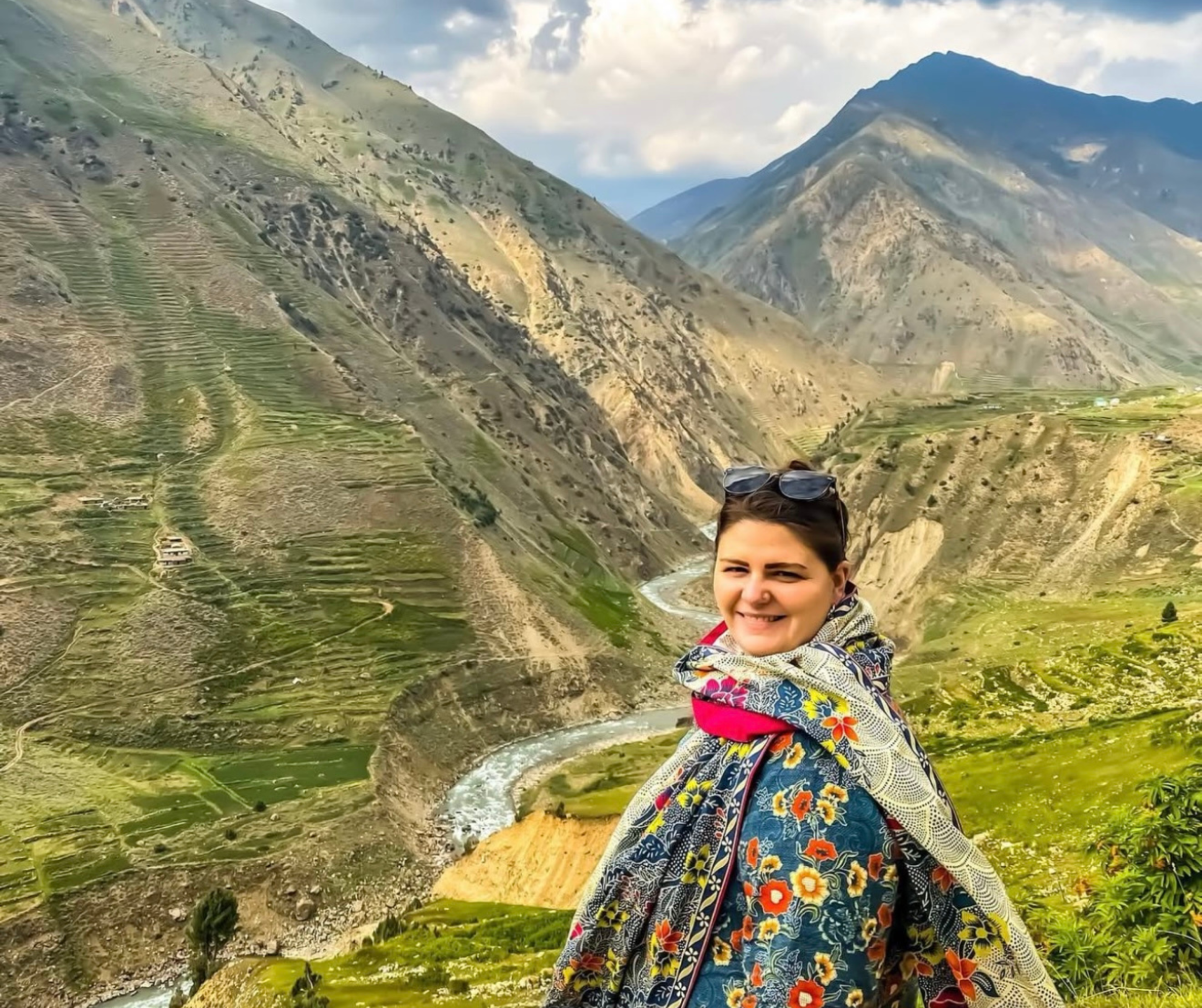 A person wearing a colorful floral shawl stands smiling in front of rolling green mountains and a winding river under a cloudy blue sky.