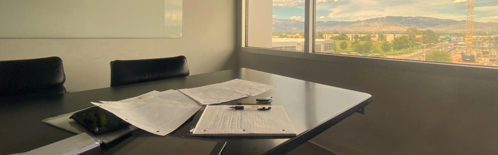 An office features a black table with scattered papers and pens. A large window shows a scenic view of mountains under a partly cloudy sky.