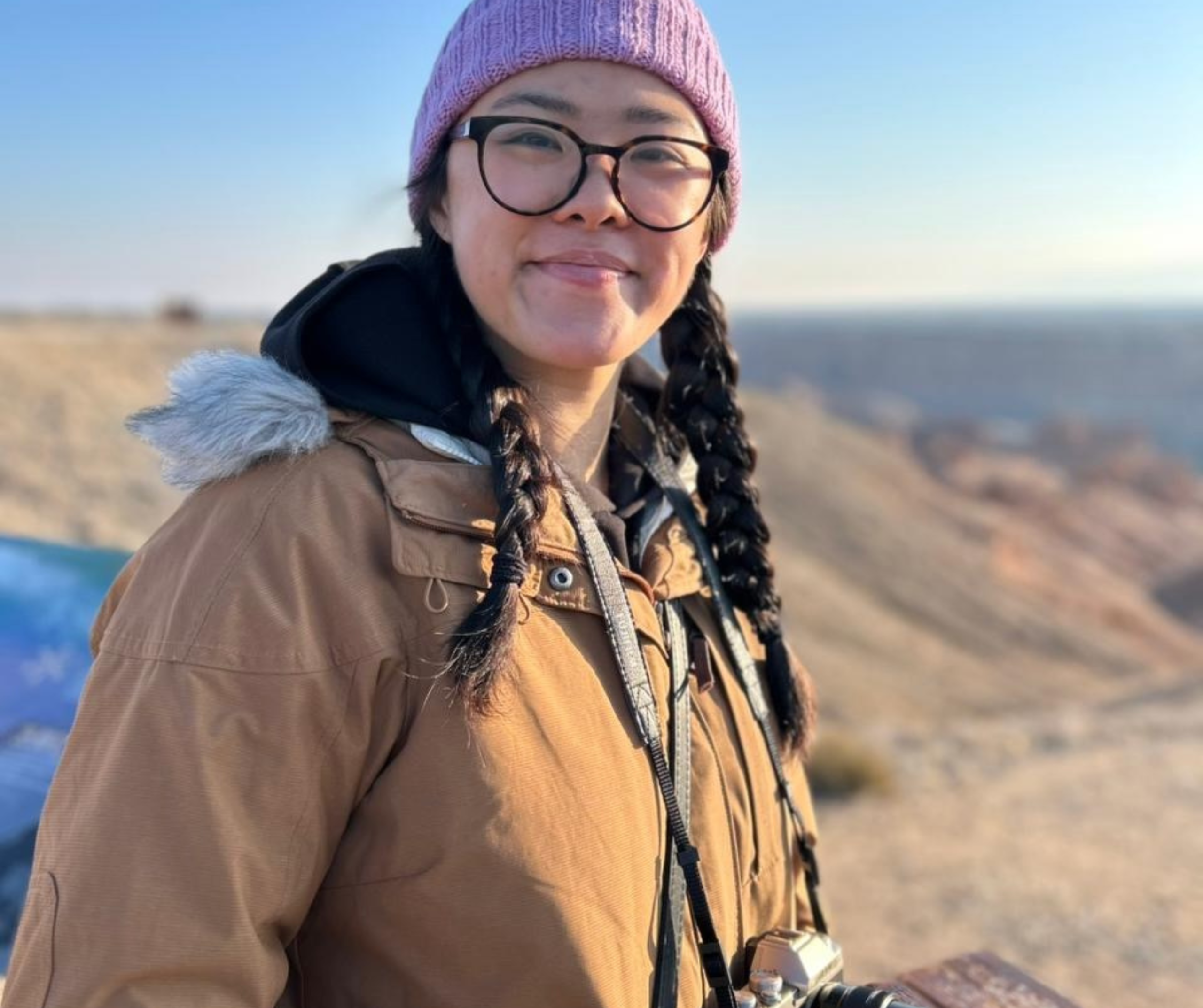 A person with glasses, stocking hat, and braids smiling with rolling hills in the background.