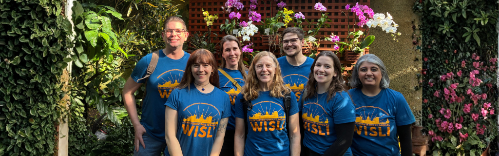 Seven smiling adults in matching blue “WISLI” T-shirts pose together in a bright greenhouse filled with lush greenery and blooming orchids.