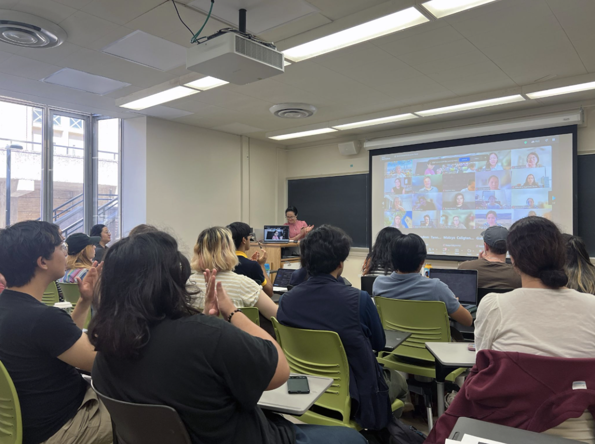 A classroom full of seated attendees applauds as a presenter speaks at the front beside a large projection screen showing a video call with multiple remote participants.