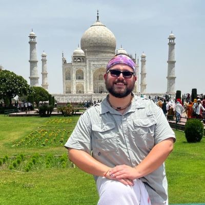 Joe stands smiling in front of the Taj Mahal in Agra, India, wearing sunglasses, a headscarf, and a light gray shirt, with the monument and its gardens visible behind him.