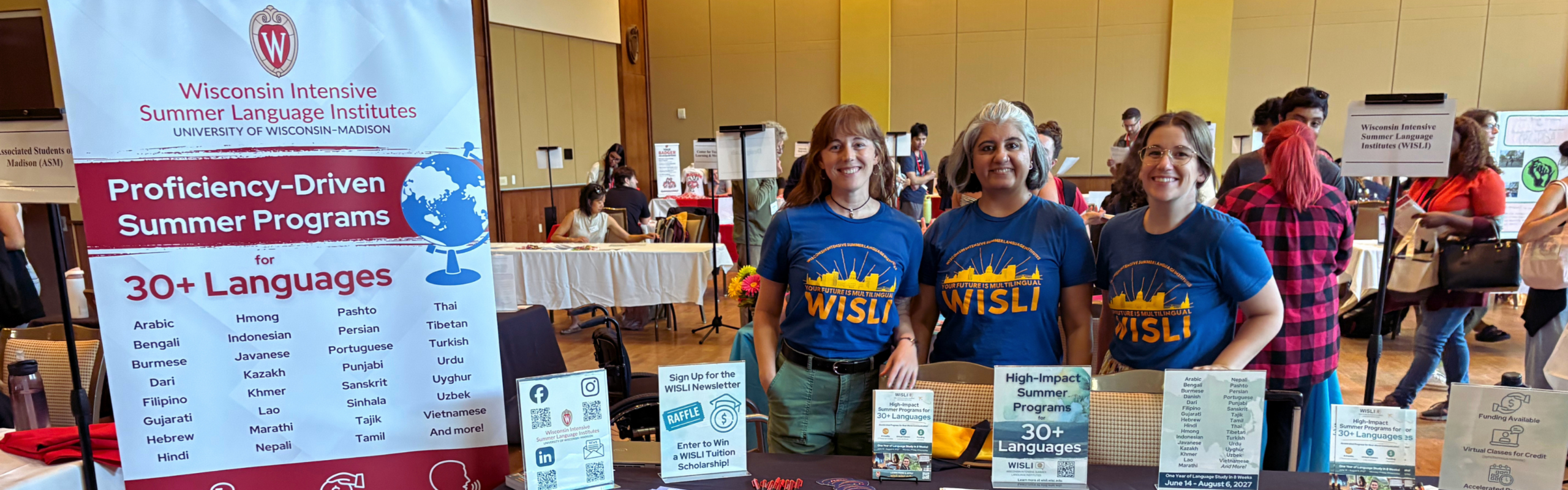 Three WISLI staff members stand behind an information table promoting proficiency-driven summer programs in 30+ languages at a campus resource fair.